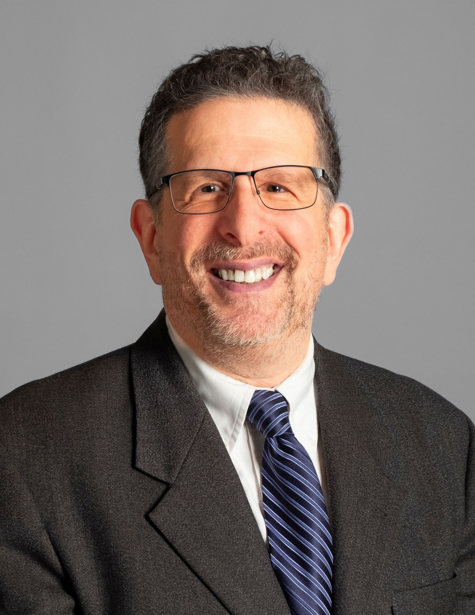 This image is a professional headshot of a man with short, dark hair. He is wearing glasses, a dark suit jacket, a white dress shirt, and a blue striped tie. The background is a simple gray, and he is smiling.