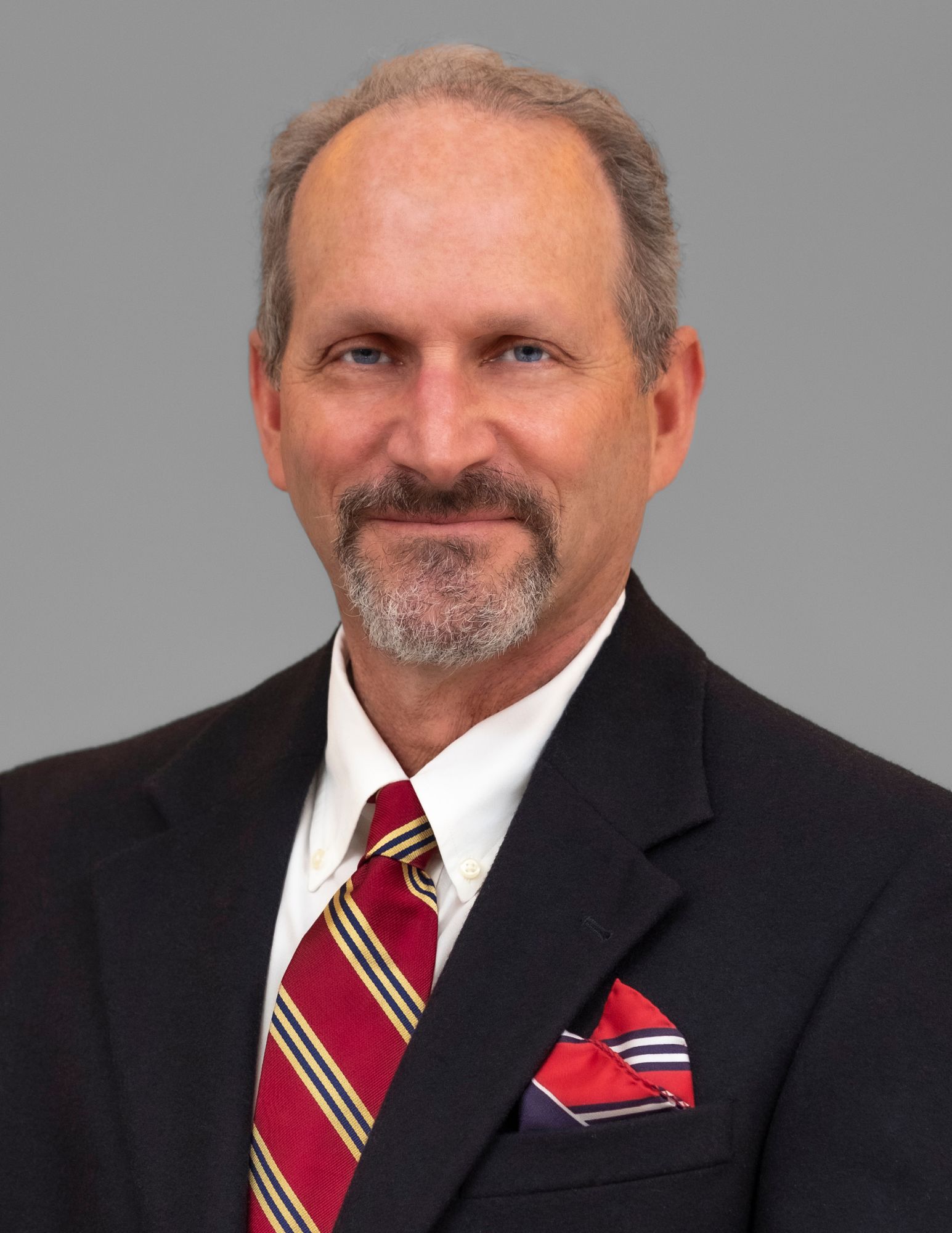This image shows a man with short, gray hair with a beard. He is wearing a black suit jacket, a white dress shirt, and a red patterned tie. He is smiling and is posing against a plain gray background.
