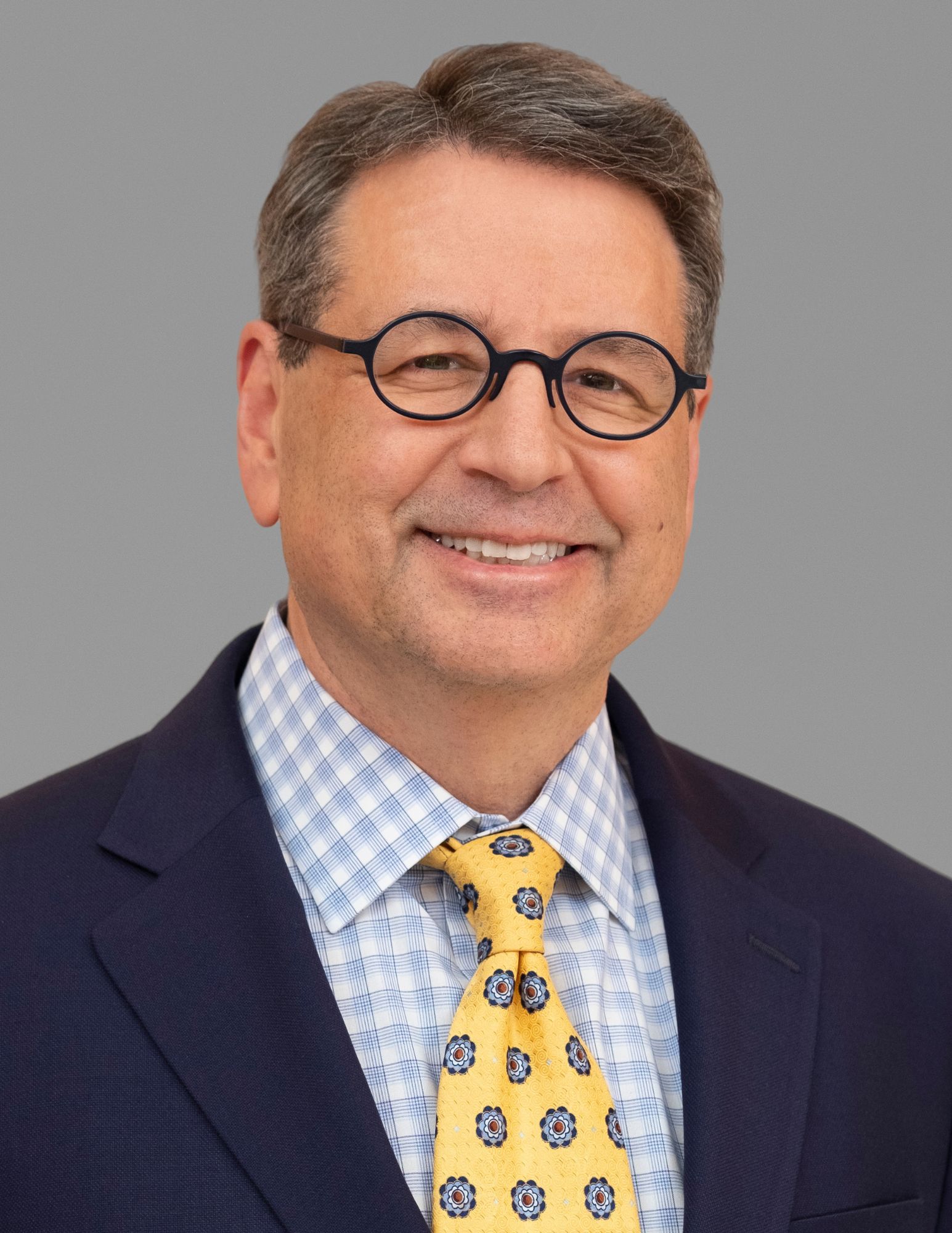This image shows a man with short, dark hair wearing round glasses, a dark blue suit jacket, a blue and white patterned dress shirt, and a yellow patterned tie. He is smiling and is posing against a plain gray background.