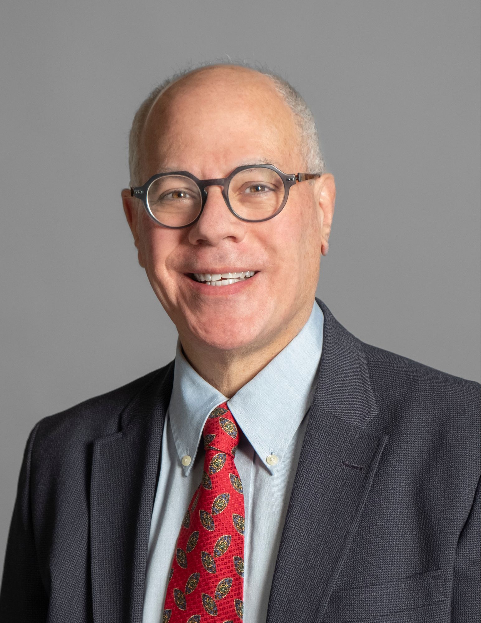 This image shows a man with short, gray hair wearing round glasses, a dark gray suit jacket, a blue dress shirt, and a red patterned tie. He is smiling and is posing against a plain gray background.