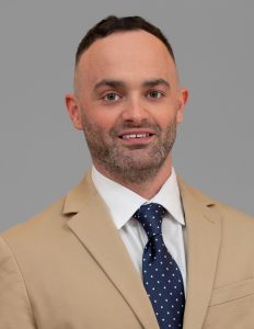 This image shows a man with short, dark hair wearing a tan suit jacket, a white dress shirt, and a blue and white polka dot tie. He is smiling and is posing against a plain gray background.