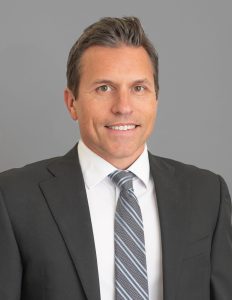 This image shows a man with short, dark hair wearing a black suit jacket, a white dress shirt, and a gray and white striped tie. He is smiling and is posing against a plain gray background.
