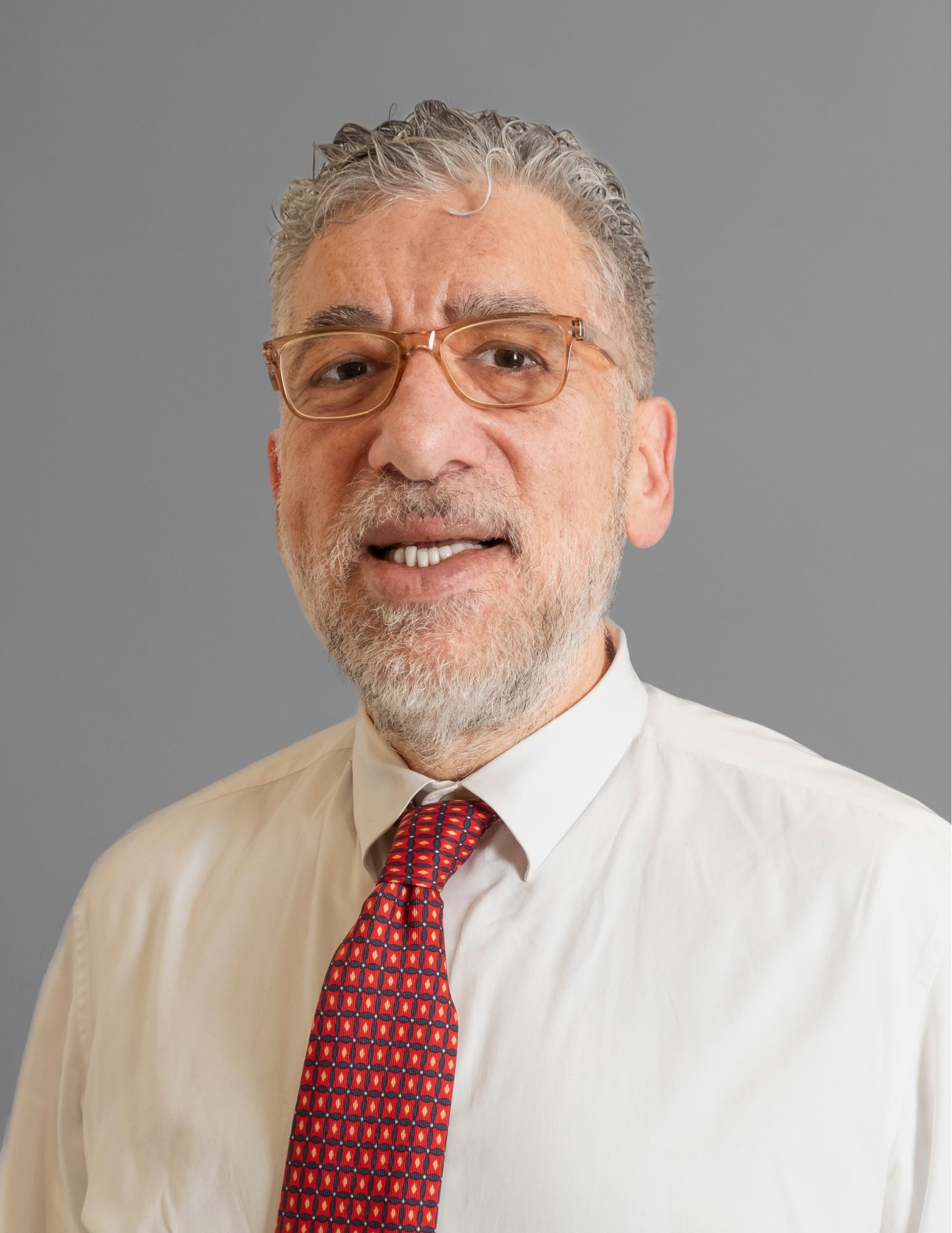 This image shows a man with short, gray curly hair and a beard. He is wearing glasses, a white dress shirt, and a red patterned tie. He is smiling slightly and is posing against a plain gray background.